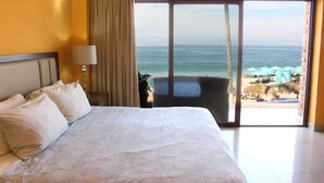 Habitación de hotel con una cama grande, una mesita de noche, una lámpara y vistas al océano y la playa a través de una puerta corrediza de cristal.