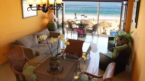 Una habitación con una mesa de vidrio, sillas de mimbre, un sofá y vista a la playa.