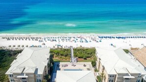 Un hotel frente a la playa con varias construcciones, una piscina y una playa con sombrillas y gente.