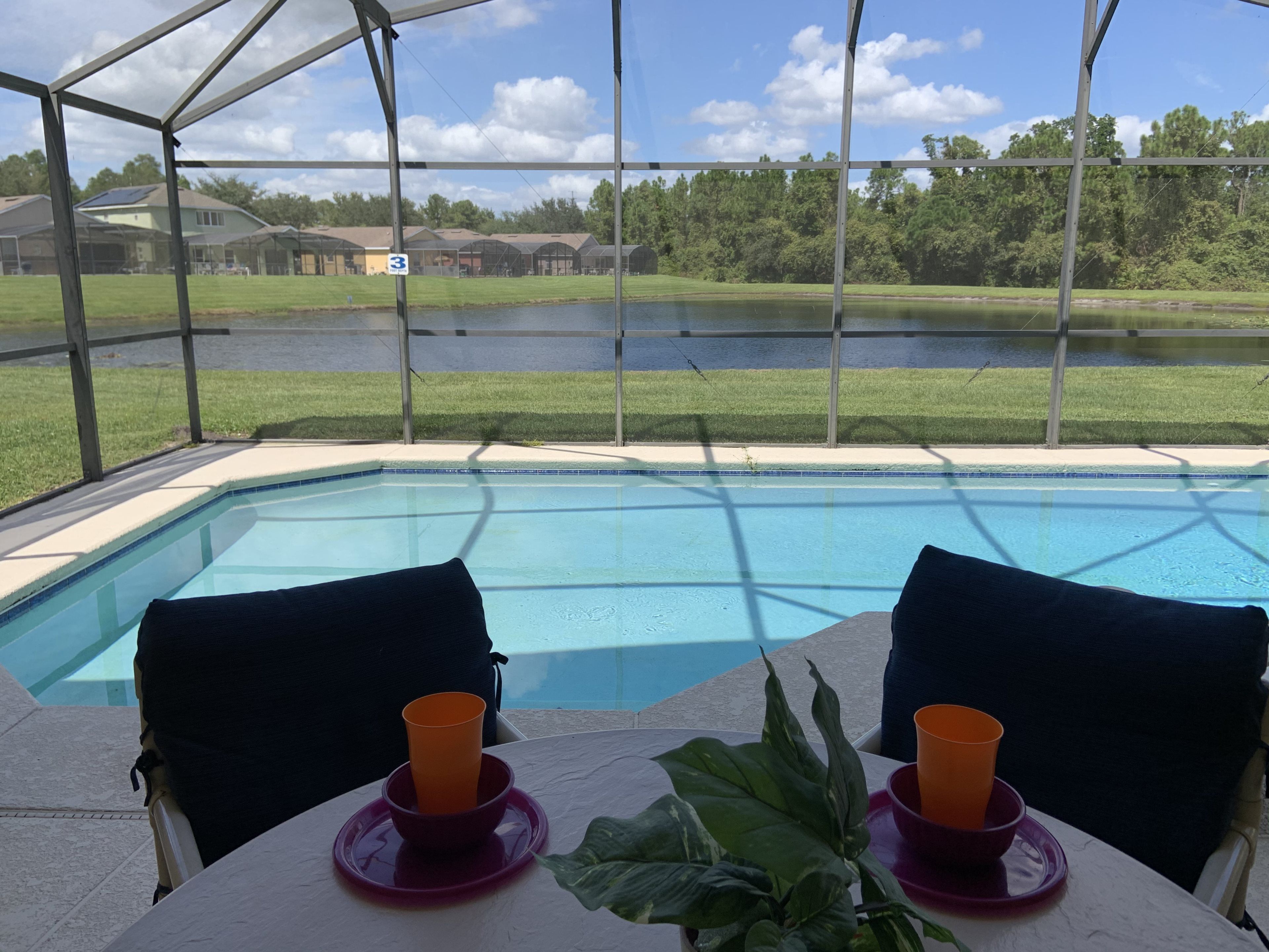 Un espace piscine doté d’une partie couverte, de deux chaises avec des coussins et d’une table sur laquelle se trouvent deux tasses oranges et un bol.