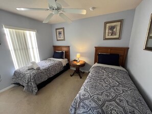 A bedroom with two beds, a ceiling fan, a nightstand with a lamp, and framed pictures on the walls.