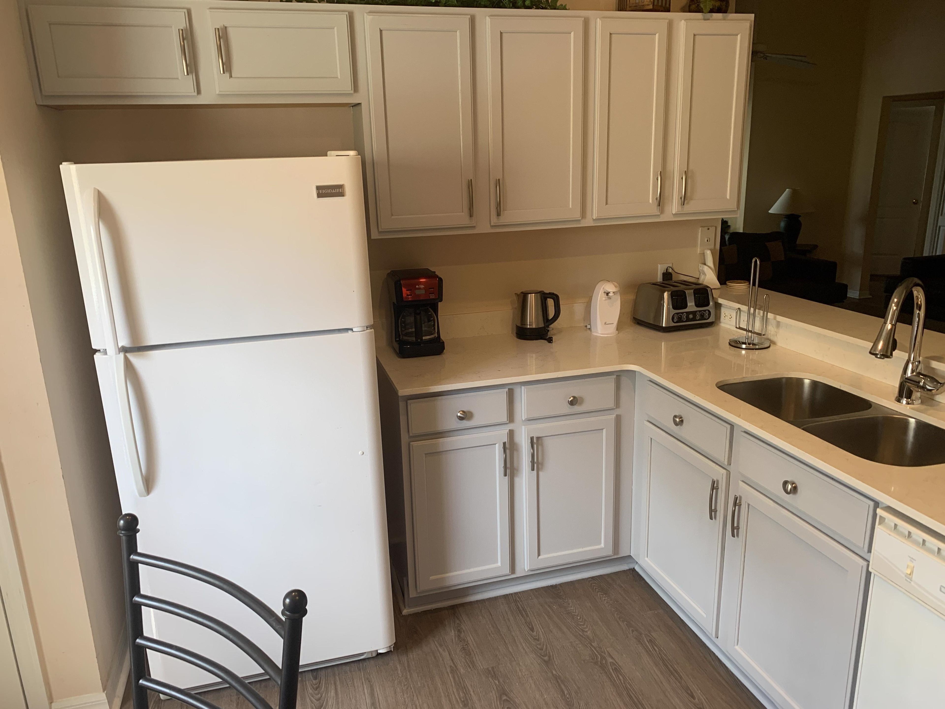 Una cocina con armarios blancos, un refrigerador blanco, una cafetera, un tostador y un lavamanos.