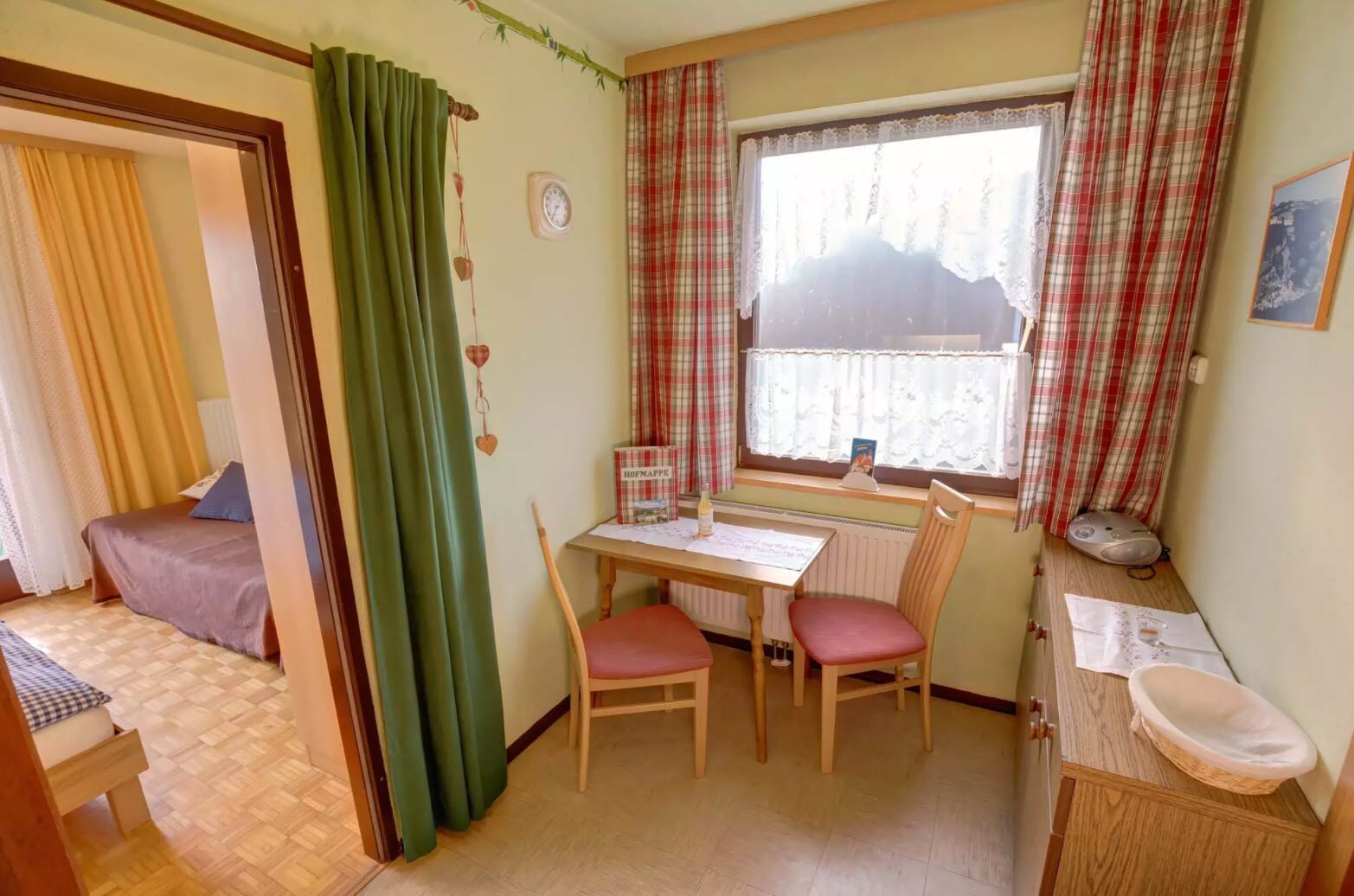 Een knus ingerichte kamer met een bed, een tafel met stoelen, een raam met gordijnen, een radiator en een klein wastafeltje.