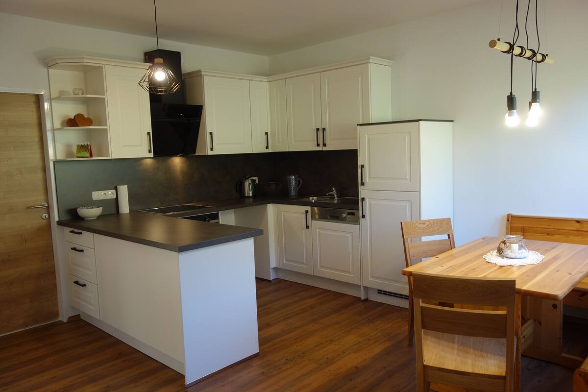 Una cocina compacta con armarios blancos, una mesa de comedor de madera y una lámpara colgante.