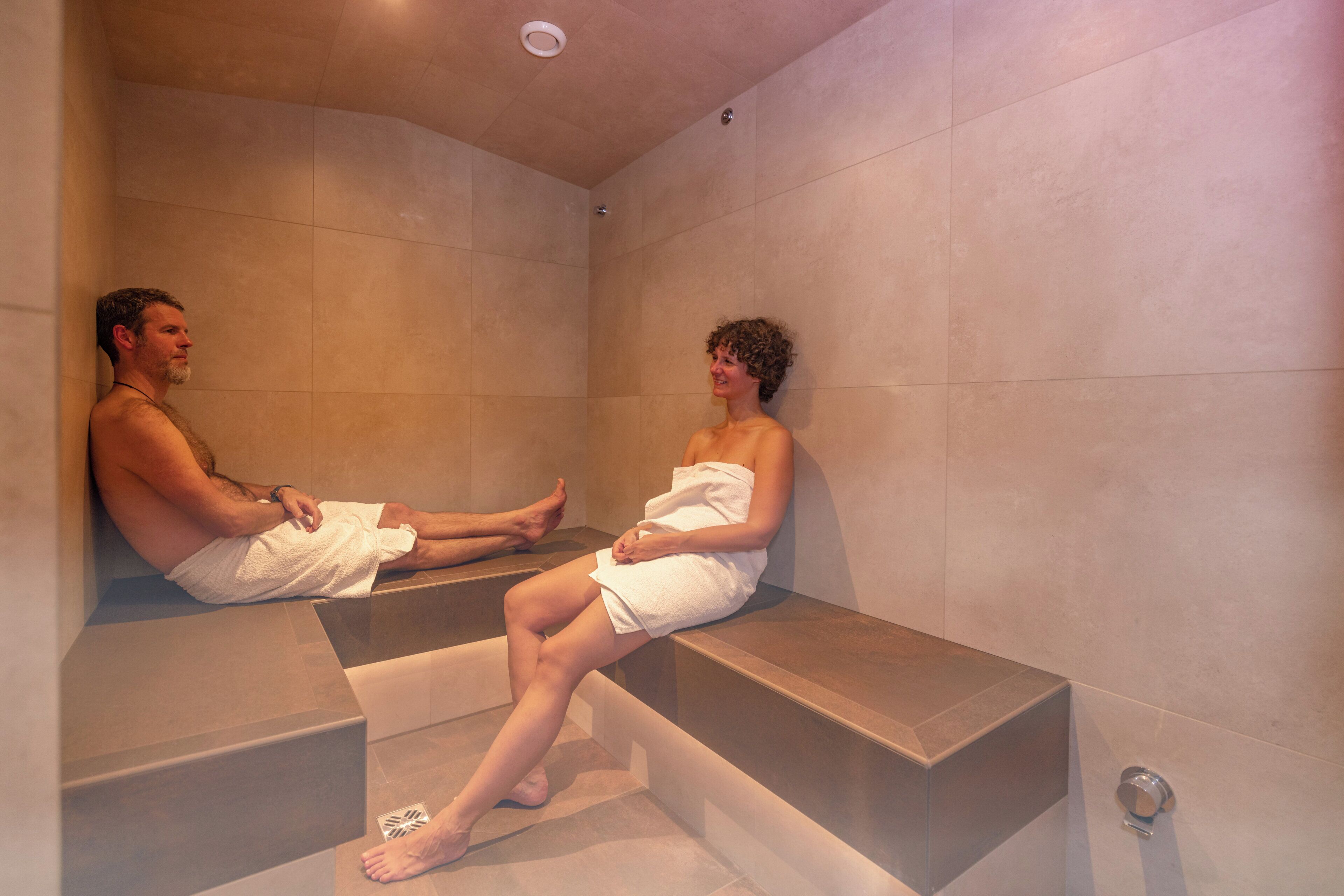 Una coppia seduta su una panchina in un ambiente simile a una spa, con pareti piastrellate e una luce a soffitto.