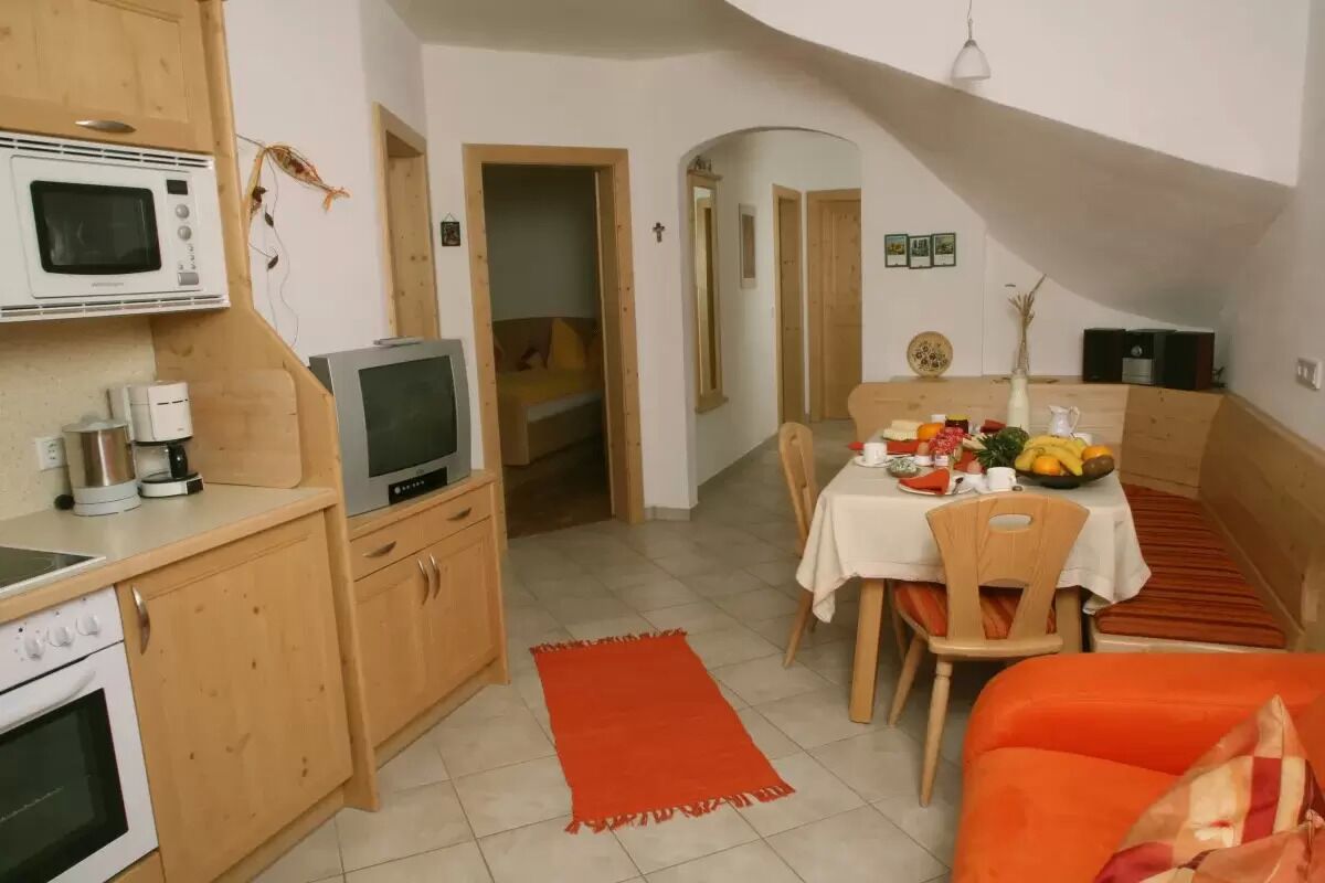 A compact kitchen with a microwave, stove, and a small TV. There's a dining area with a table set for two, a red rug, and a small seating area with an orange couch.