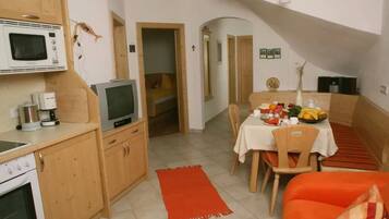 A compact kitchen with a microwave, stove, and a small TV. There's a dining area with a table set for two, a red rug, and a small seating area with an orange couch.