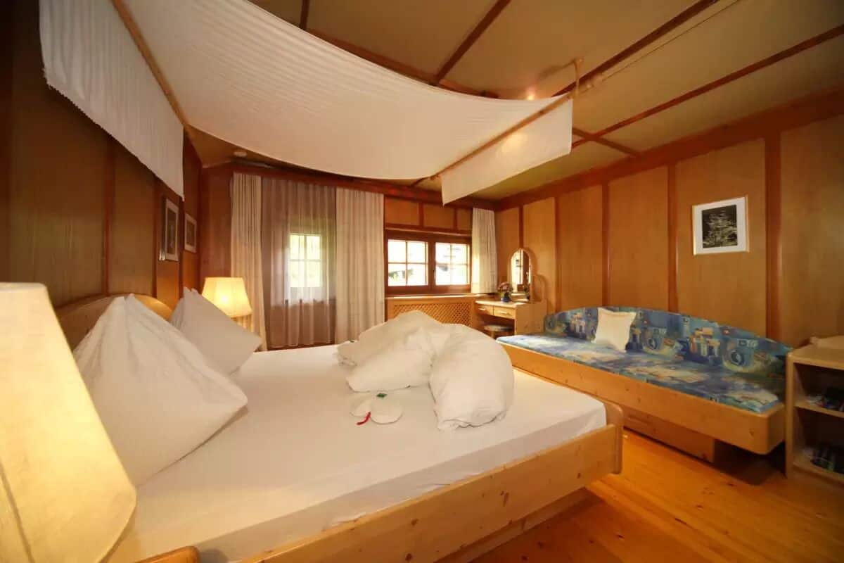 Una habitación con una cama de madera, una cama individual con colchón azul, un escritorio con lámpara y una ventana con cortinas.