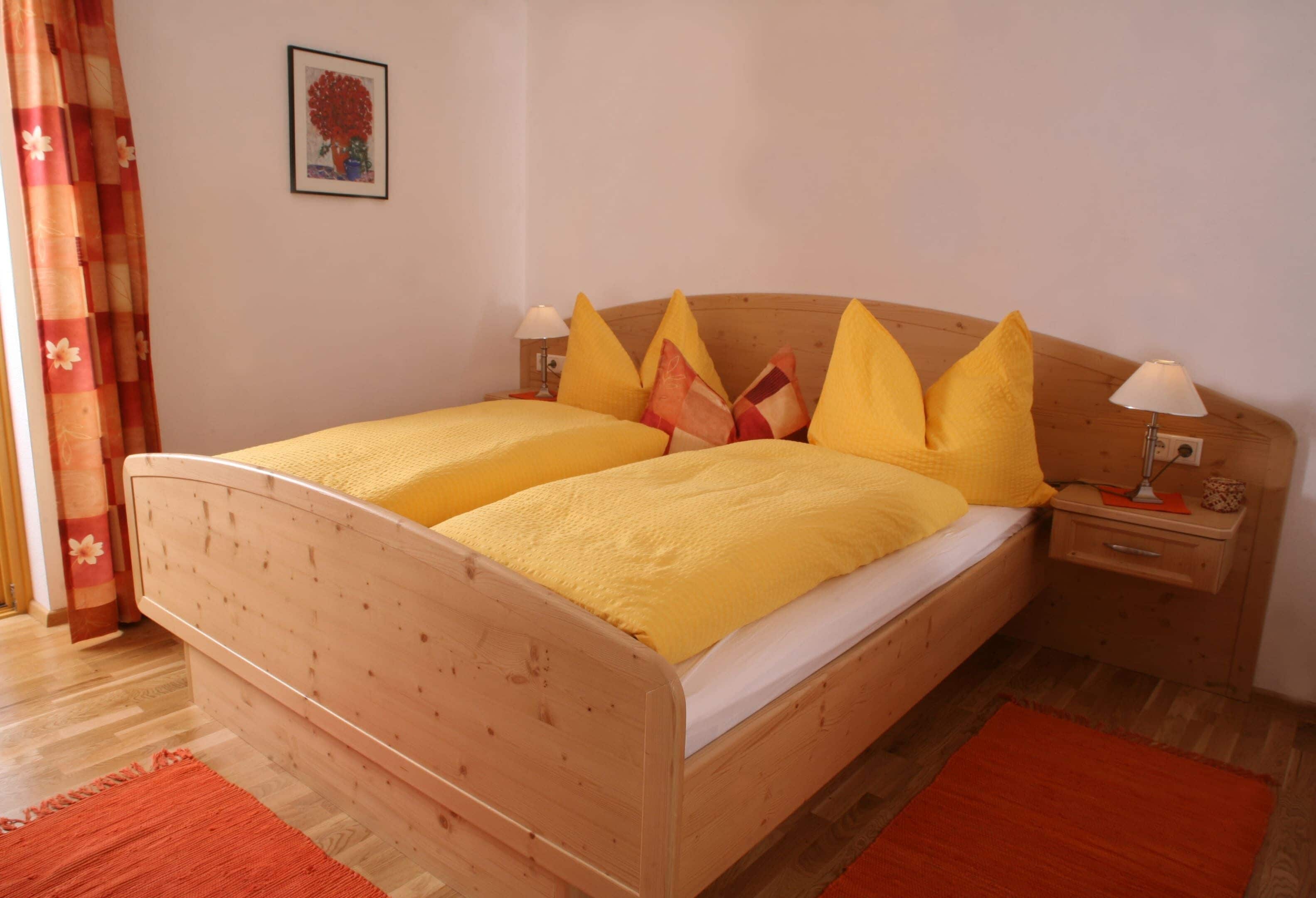 Una cama doble con ropa de cama amarilla, dos mesitas de noche con lámparas, un cabecero de madera, una alfombra roja y un cuadro enmarcado en la pared.
