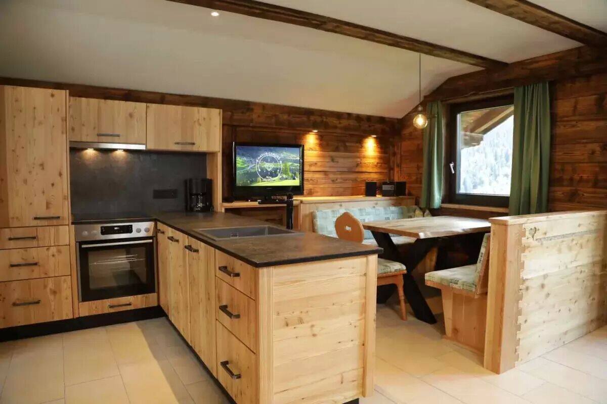Una cocina compacta con muebles de madera, horno empotrado, zona de comedor con banco y televisor montado en la pared.