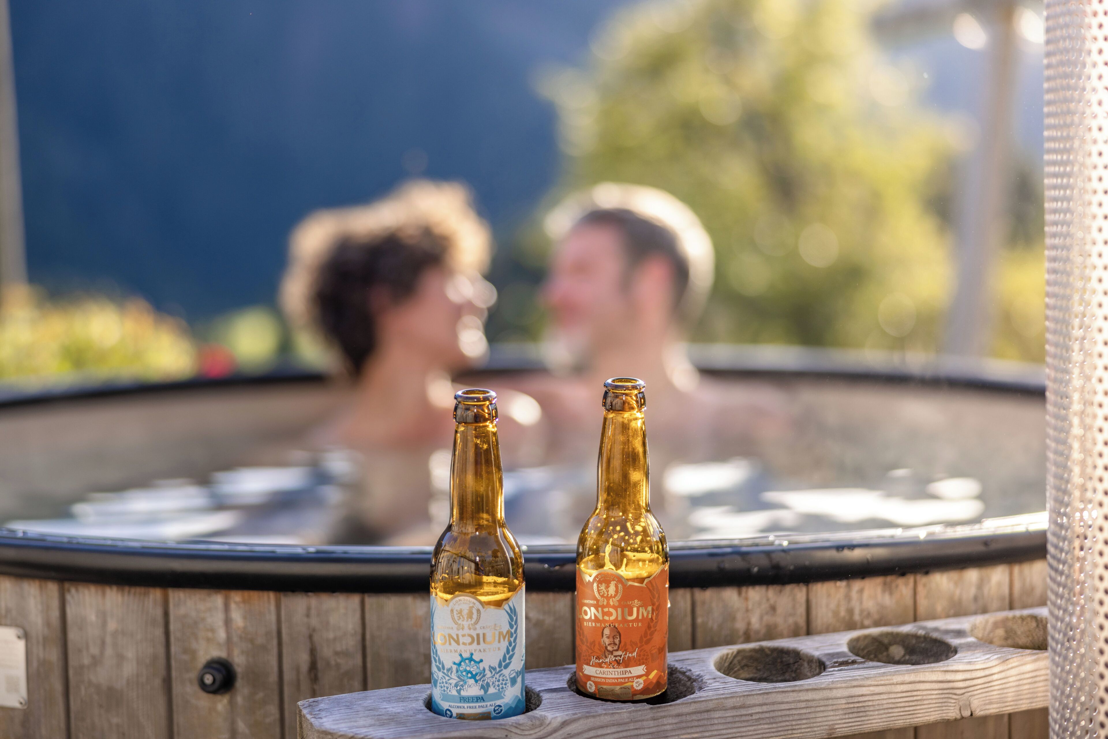 Una vasca idromassaggio con due bottiglie di birra, panche di legno e sullo sfondo figure sfocate.