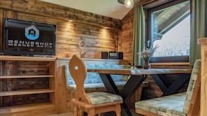 Un interior de madera con una televisión mostrando 'GENUSSHOF MESNER AKTIV', una mesa de madera con sillas y una ventana con vistas.