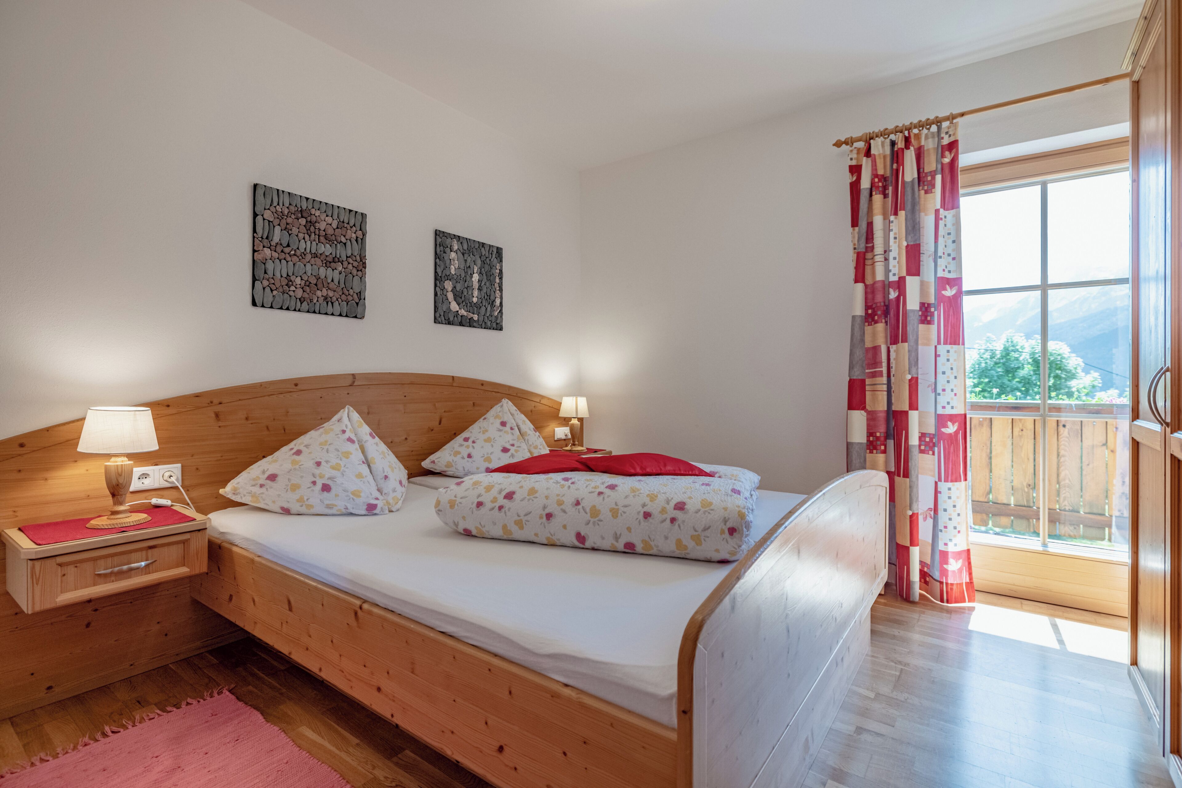 Una cama de madera con colchón blanco, almohadas y una manta roja. Hay dos mesitas de noche con lámparas y un aparador. El cuarto tiene piso de madera y un ventanal con vista a la vegetación.