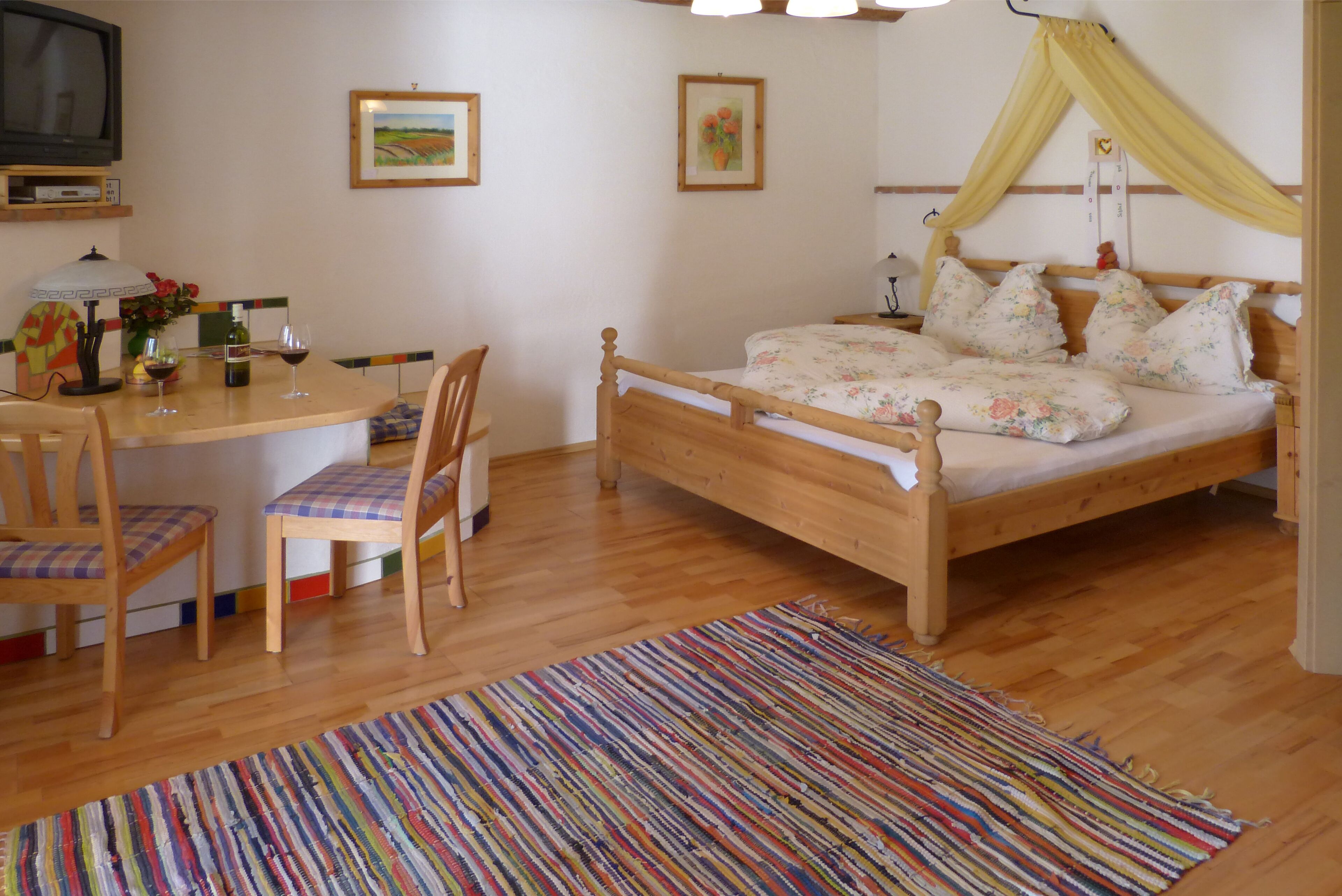 Ein Schlafzimmer mit einem Holzbett, einem Schreibtisch mit Stühlen, einem farbenfrohen Teppich und einem Fernseher an der Wand.
