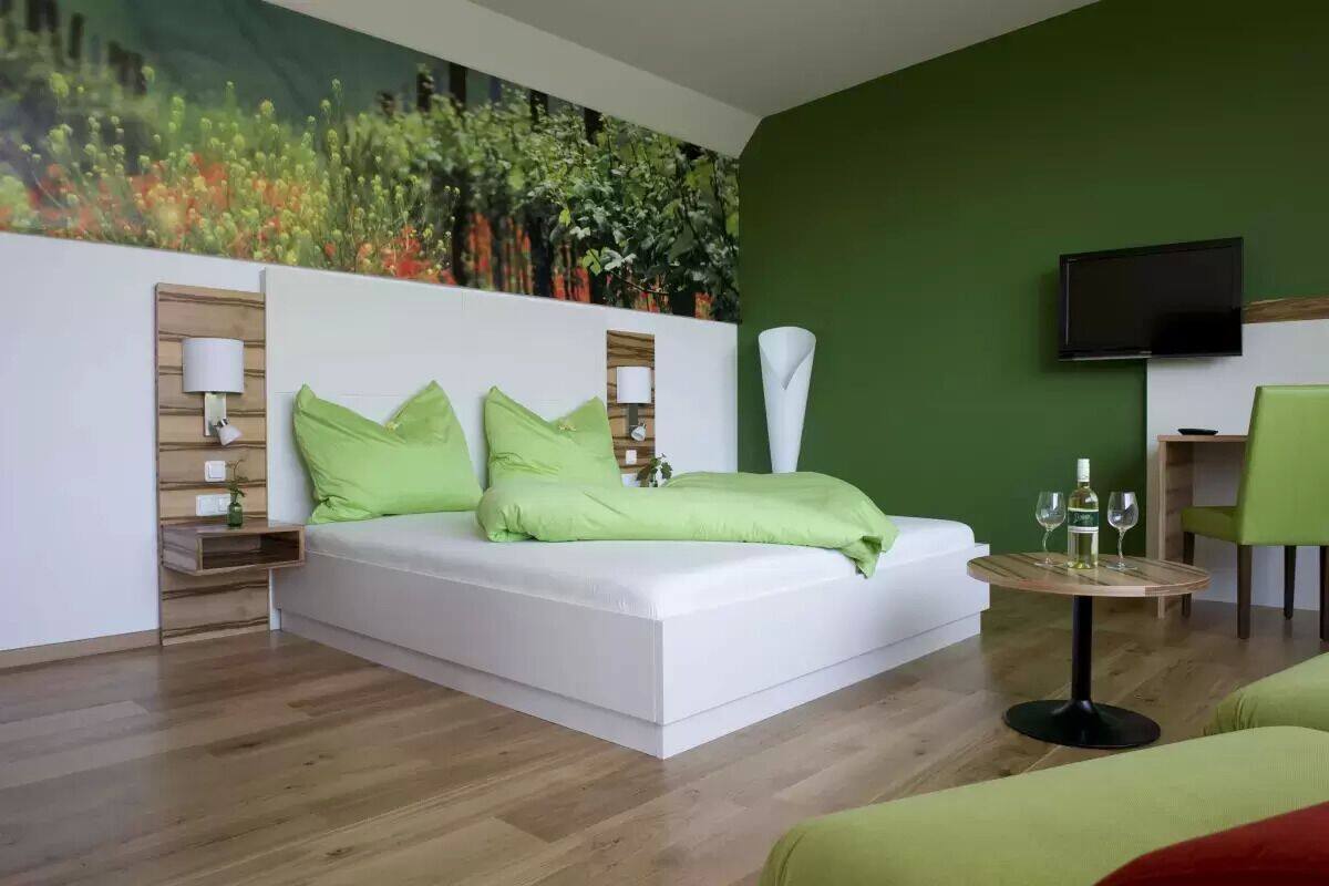 Una habitación de hotel moderna con un sofá blanco, cojines verdes, una mesa redonda de madera con vasos, un televisor de pantalla plana y una pared de acento verde.