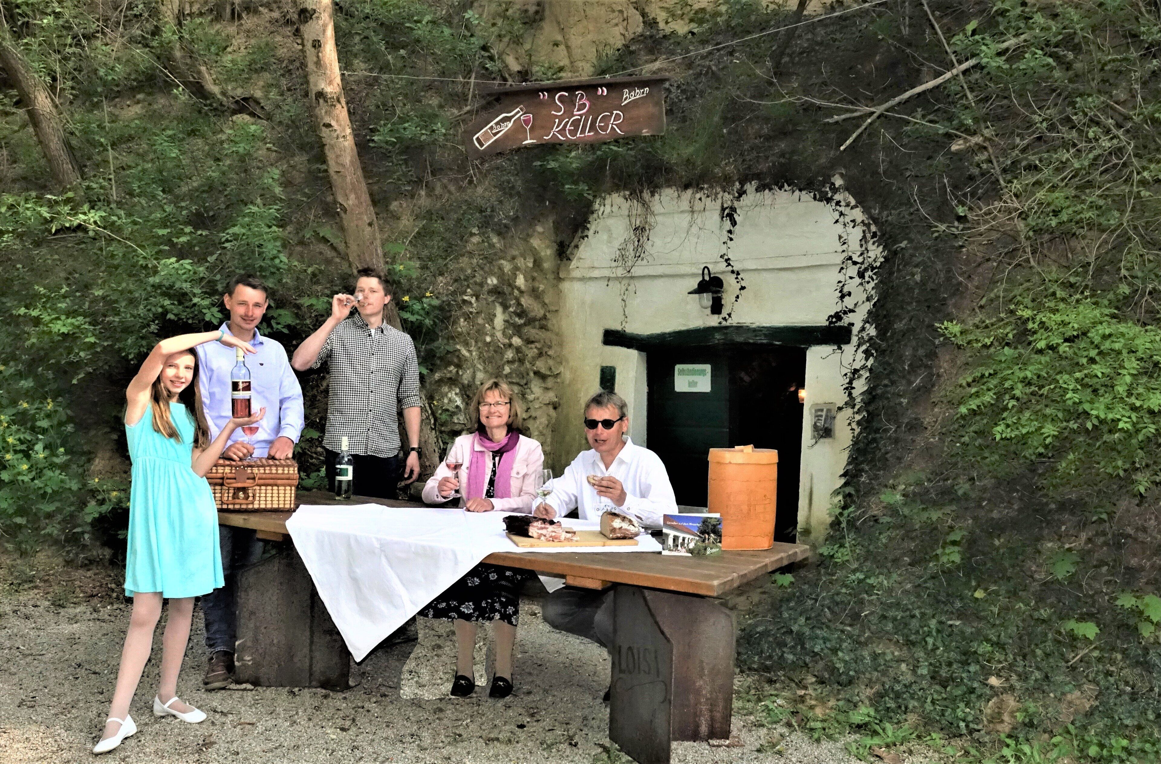 A group of people at an outdoor dining area with a rustic wooden table, surrounded by greenery.