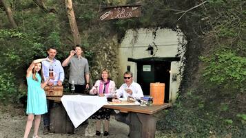 A group of people at an outdoor dining area with a rustic wooden table, surrounded by greenery.