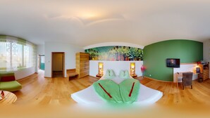 A modern hotel room with a curved ceiling, wooden flooring, a bed with green and red pillows, a desk with a chair, and a painting of a tree on the wall. - Weingut Himmelbett (Herrnbaumgarten)