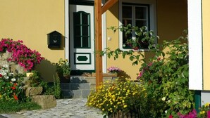 Un edificio giallo con un vialetto in pietra che conduce a una porta in legno. Sono presenti aiuole con diverse piante e fiori.