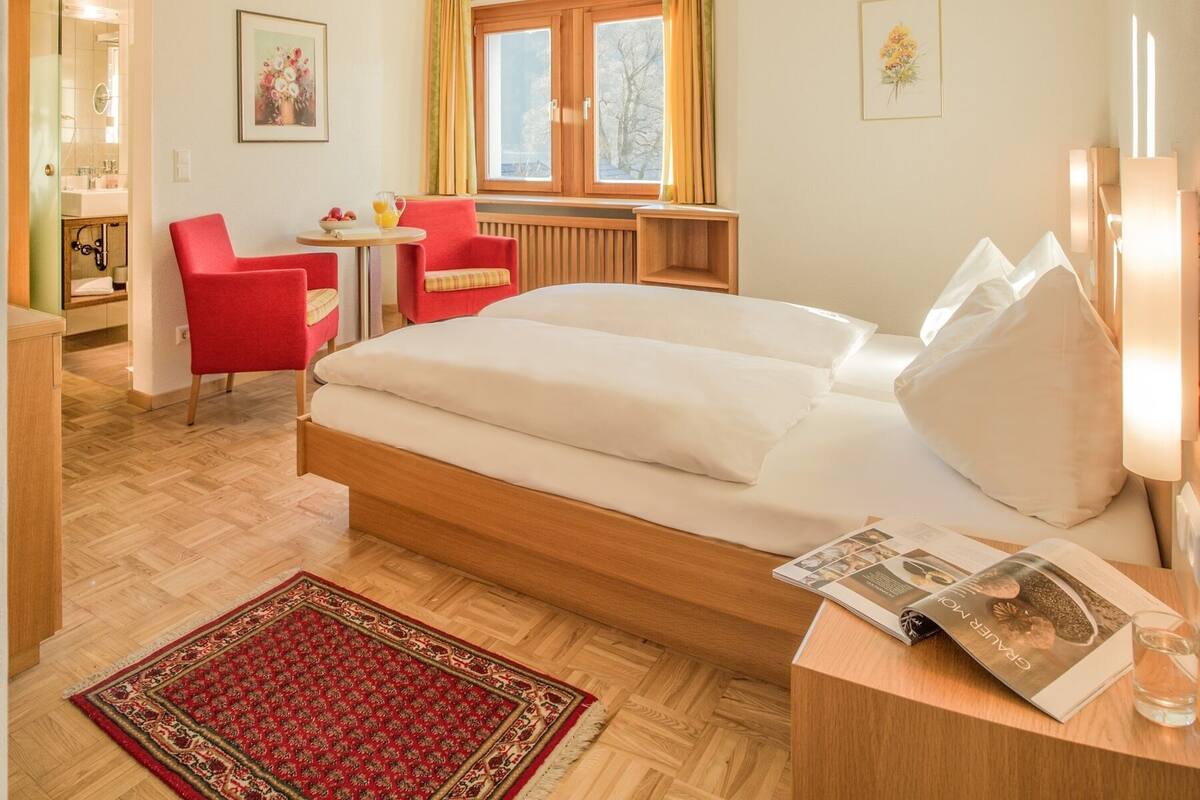 Ein Hotelzimmer mit einem großen Bett, einem kleinen Schreibtisch, einem Stuhl, einem Fenster mit Vorhängen und einem roten Teppich.