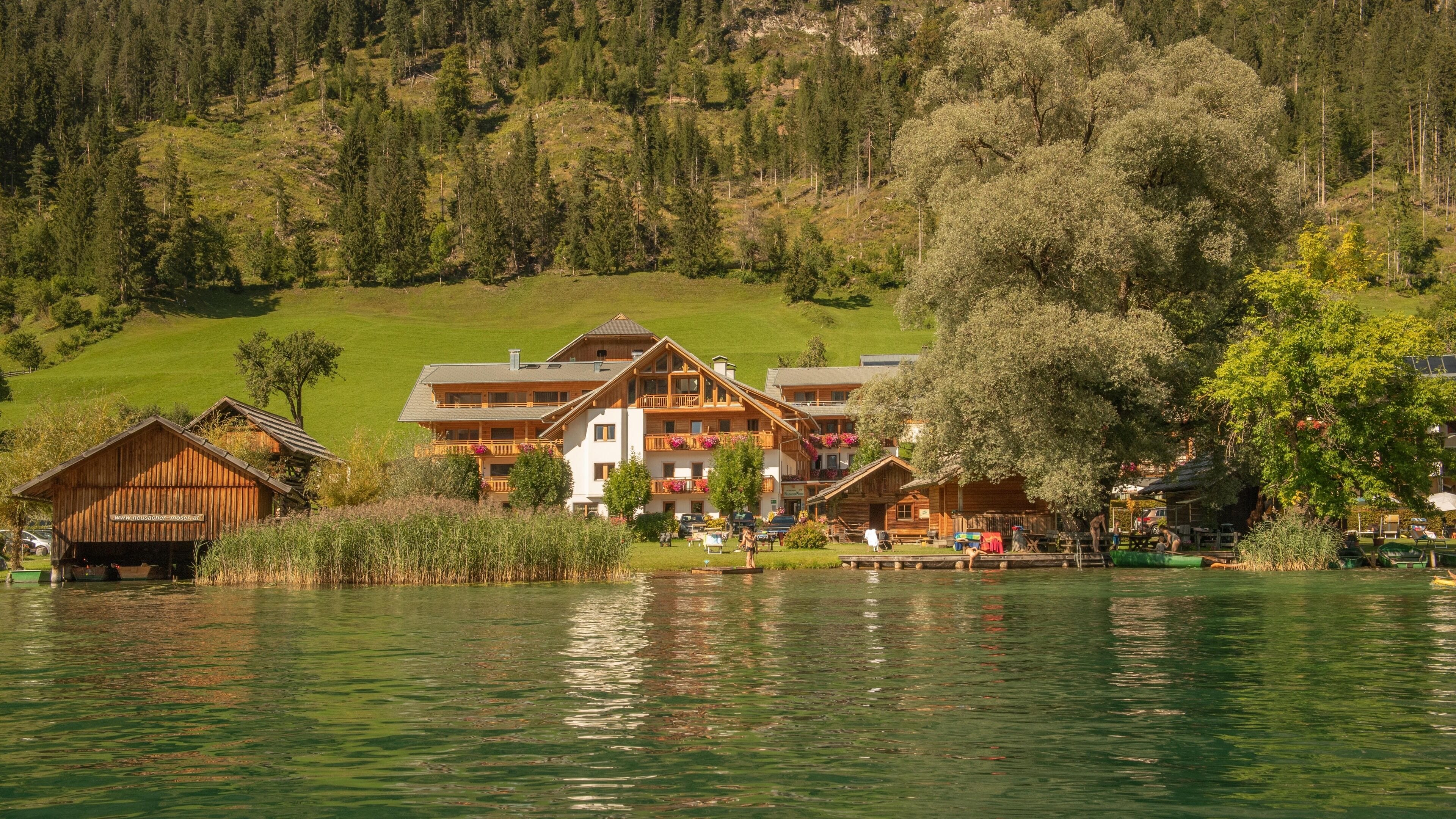 Un chalet au bord du lac, composé de plusieurs bâtiments en bois, avec un quai et une colline verdoyante en arrière-plan.