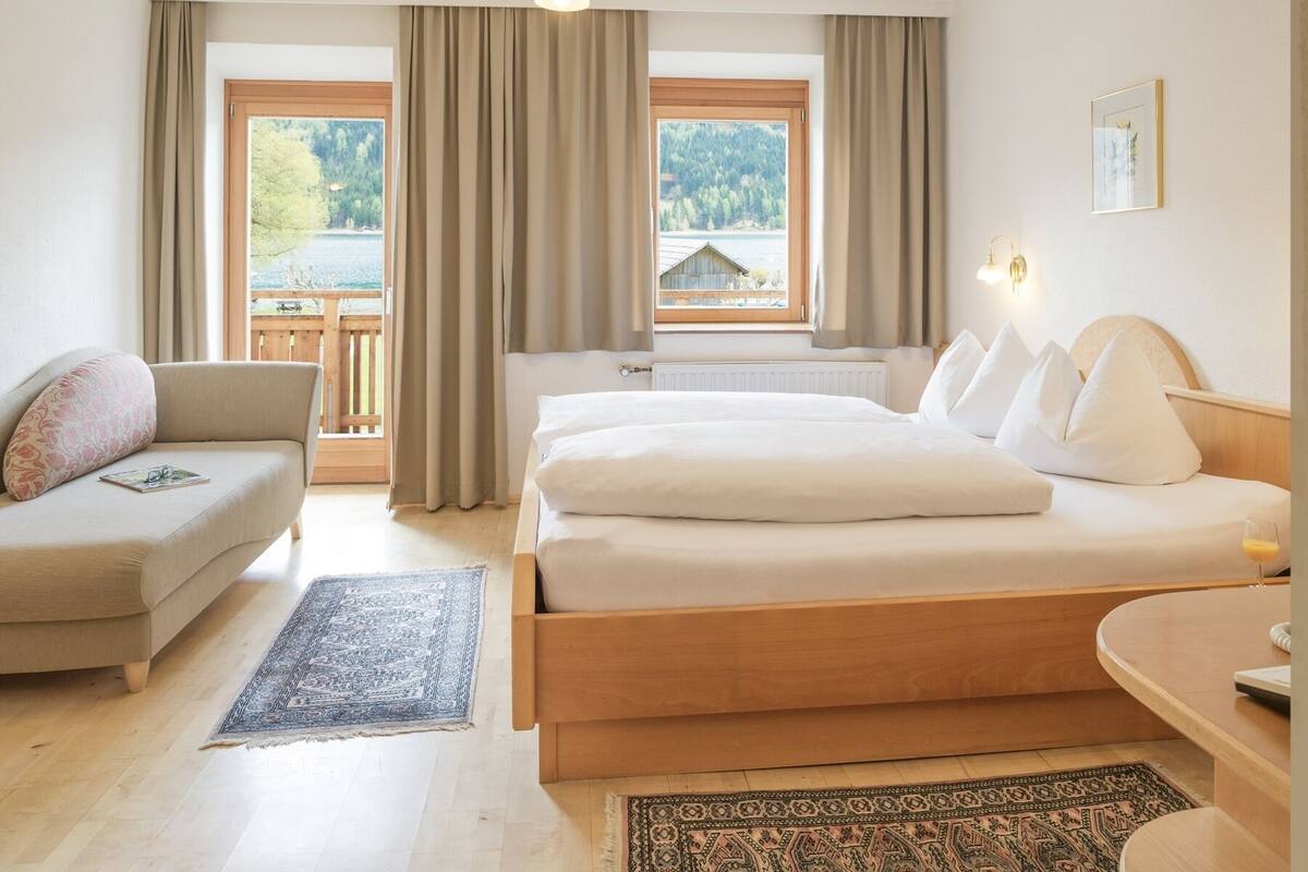 Ein Hotelzimmer mit Bett, Sofa, einem kleinen Tisch und Blick auf einen See und Wald durch große Fenster.