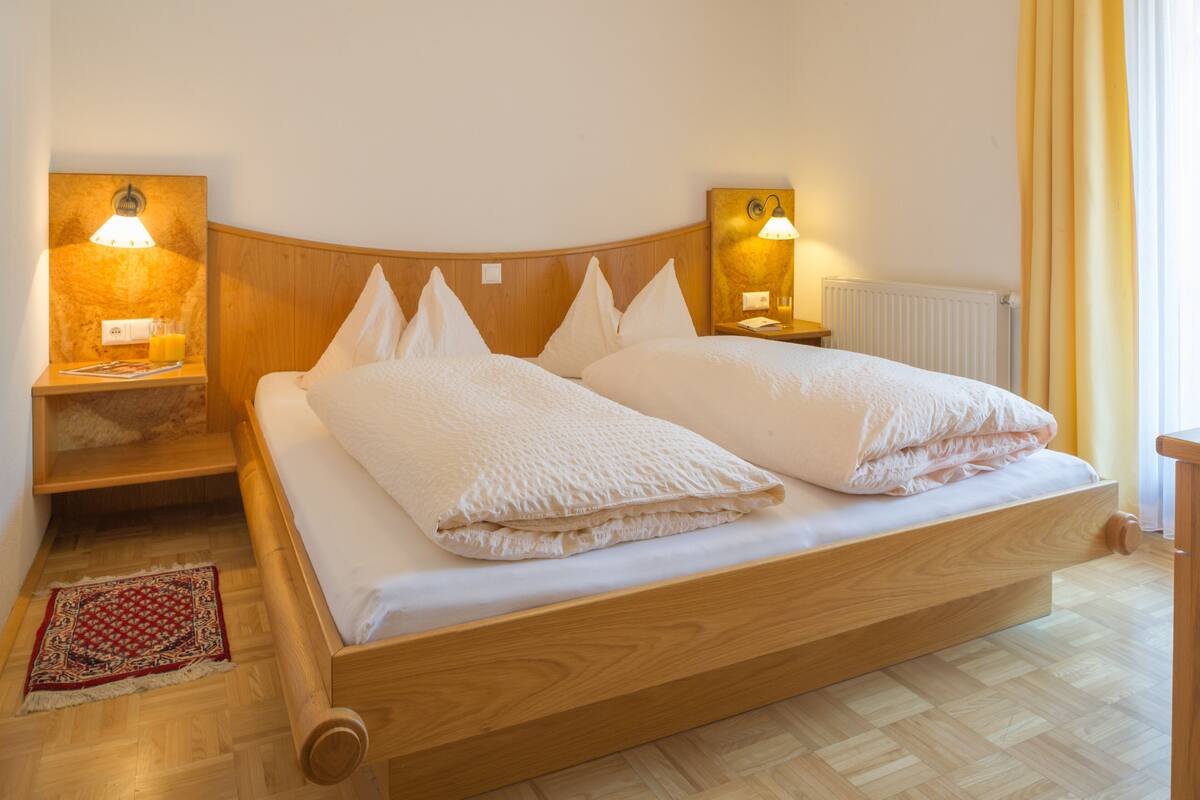 Ein Doppelbett mit Holzüberbau und zwei Nachttischen, jeder mit einer Lampe. An der Wand ist ein Heizkörper zu sehen, und rechts befindet sich ein Fenster mit Vorhängen.