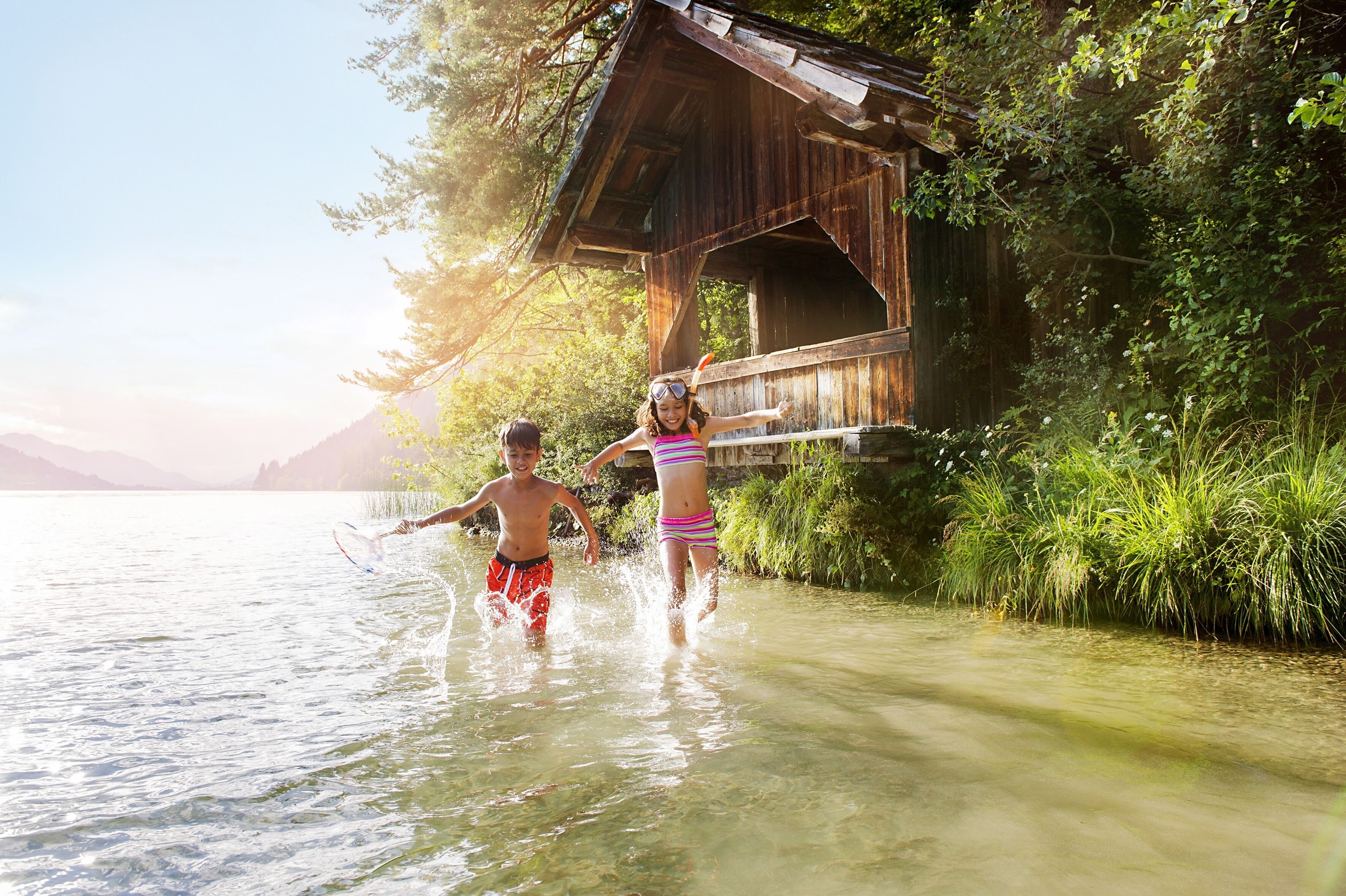 Two children playing in shallow water near a wooden cabin with a view of mountains in the background.