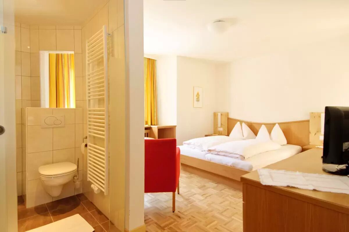 A hotel room with a wooden floor, a bed with white linens, a red chair, a wooden desk, and a bathroom with a toilet and towel rack.