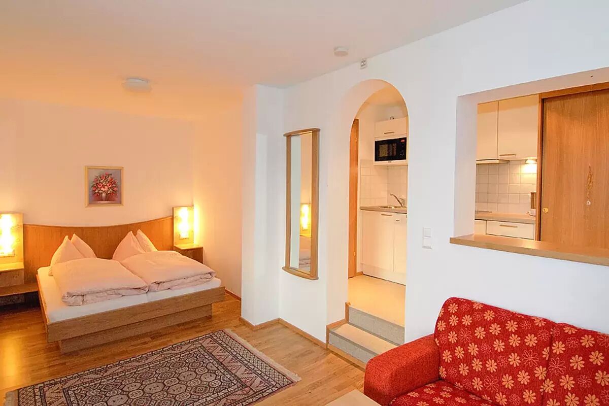 A bedroom with a bed, bedside lamps, a red sofa, a patterned rug, and a kitchen visible through an archway.