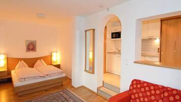 A bedroom with a bed, bedside lamps, a red sofa, a patterned rug, and a kitchen visible through an archway.