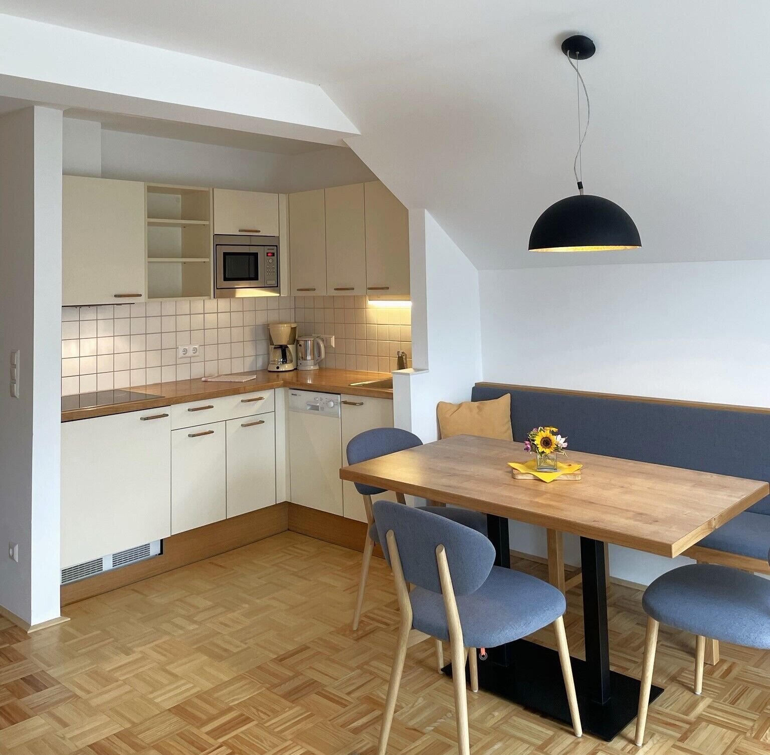 A compact kitchen with white cabinets, a microwave, a coffee maker, and a wooden dining table with blue chairs.