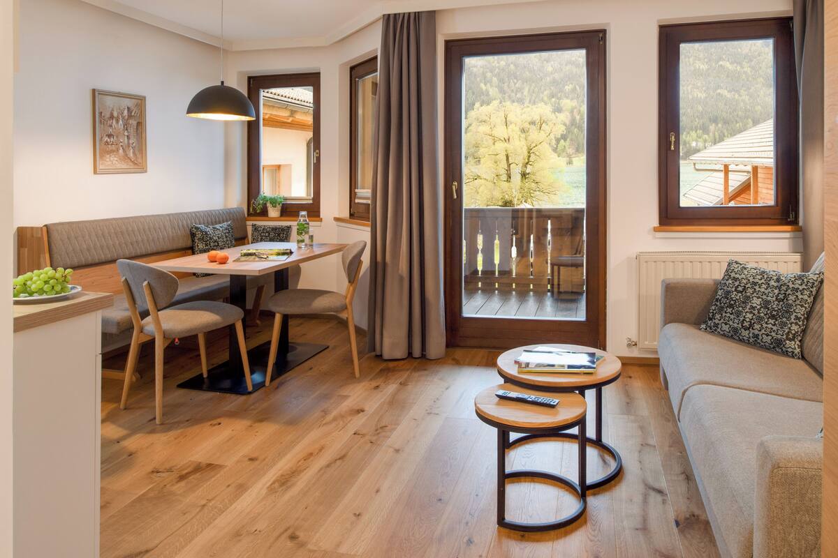 Ein gemütliches Wohnzimmer mit einer Couch, einem Esstisch mit Stühlen, einem kleinen Couchtisch und einem Balkon mit Blick auf Bäume.