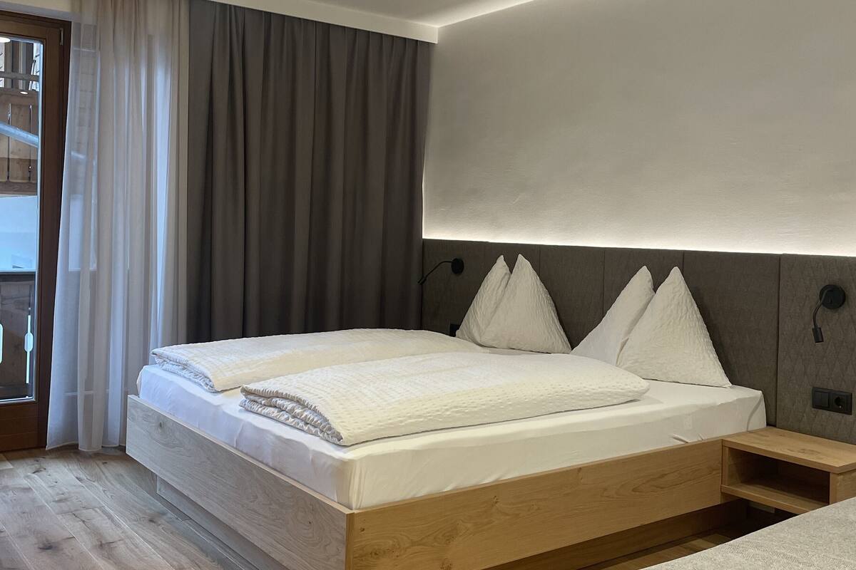 Ein modernes Schlafzimmer mit einem hölzernen Bettgestell, einem großen Fenster mit Vorhängen und einem Nachttisch.