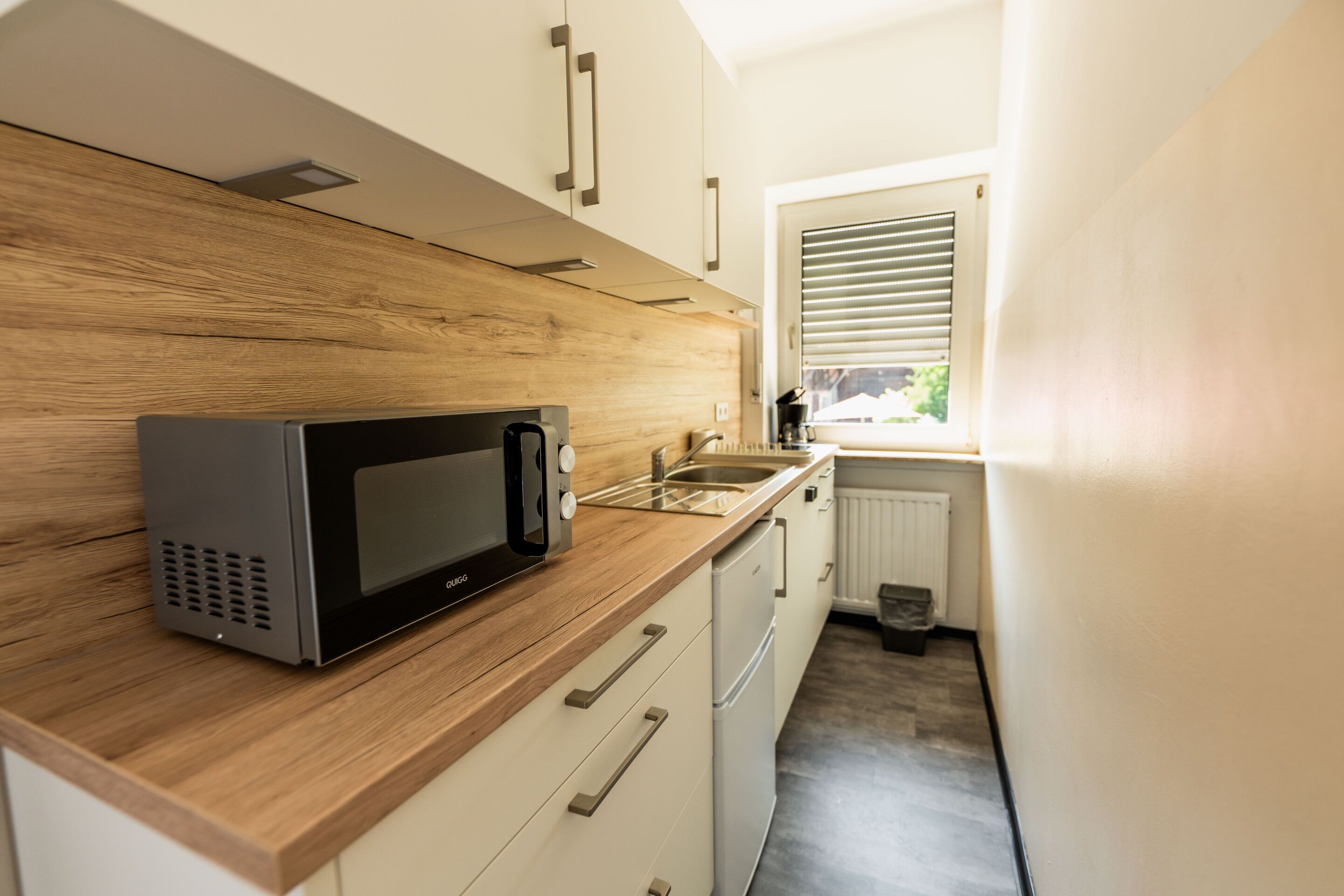 Cocina compacta con microondas, encimera de madera, armarios blancos y una ventana con persianas.