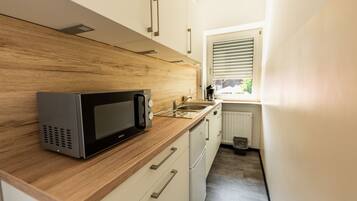 Cocina compacta con microondas, encimera de madera, armarios blancos y una ventana con persianas.