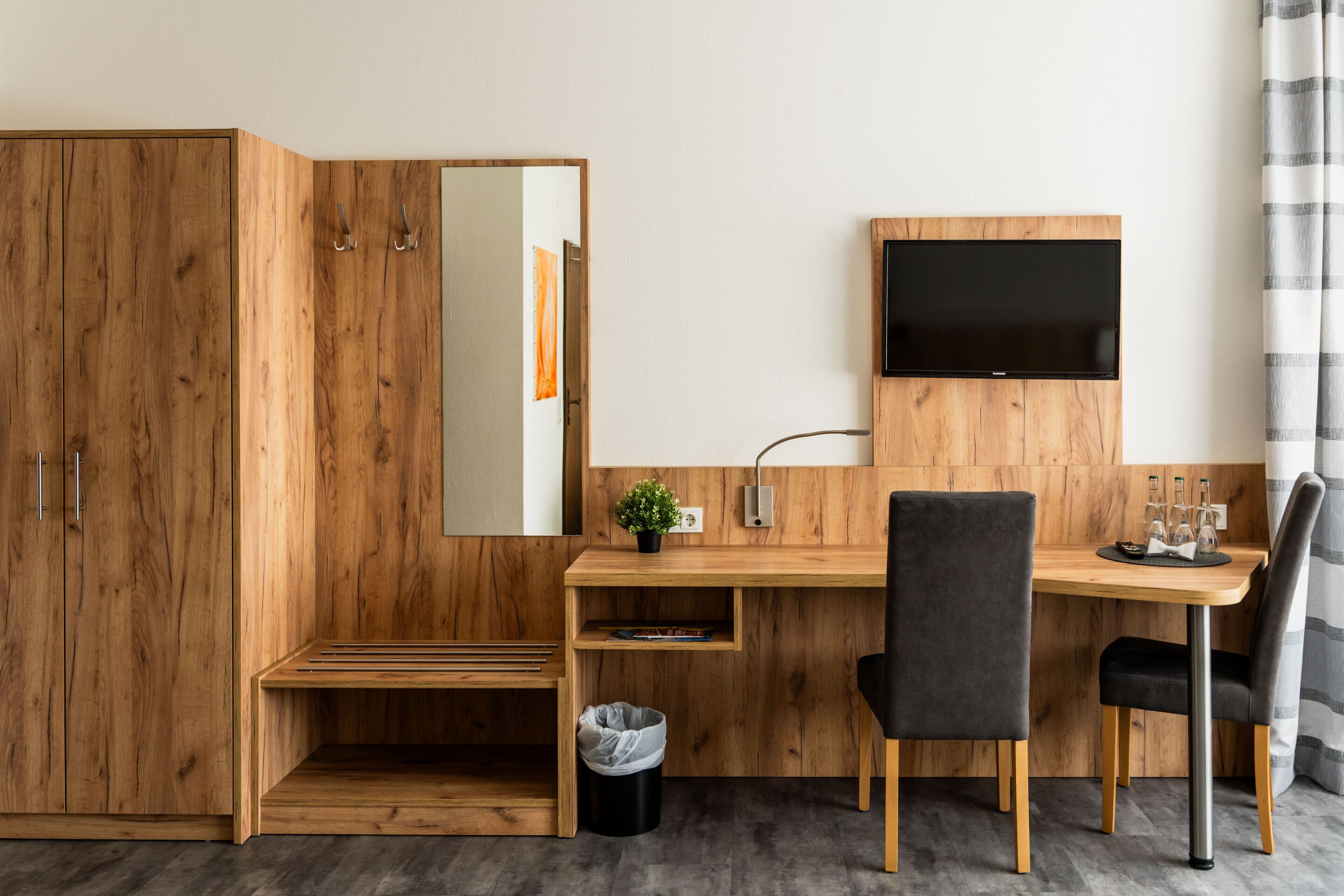 A modern room with a wooden desk, a TV mounted on the wall, a mirror, and a small plant.