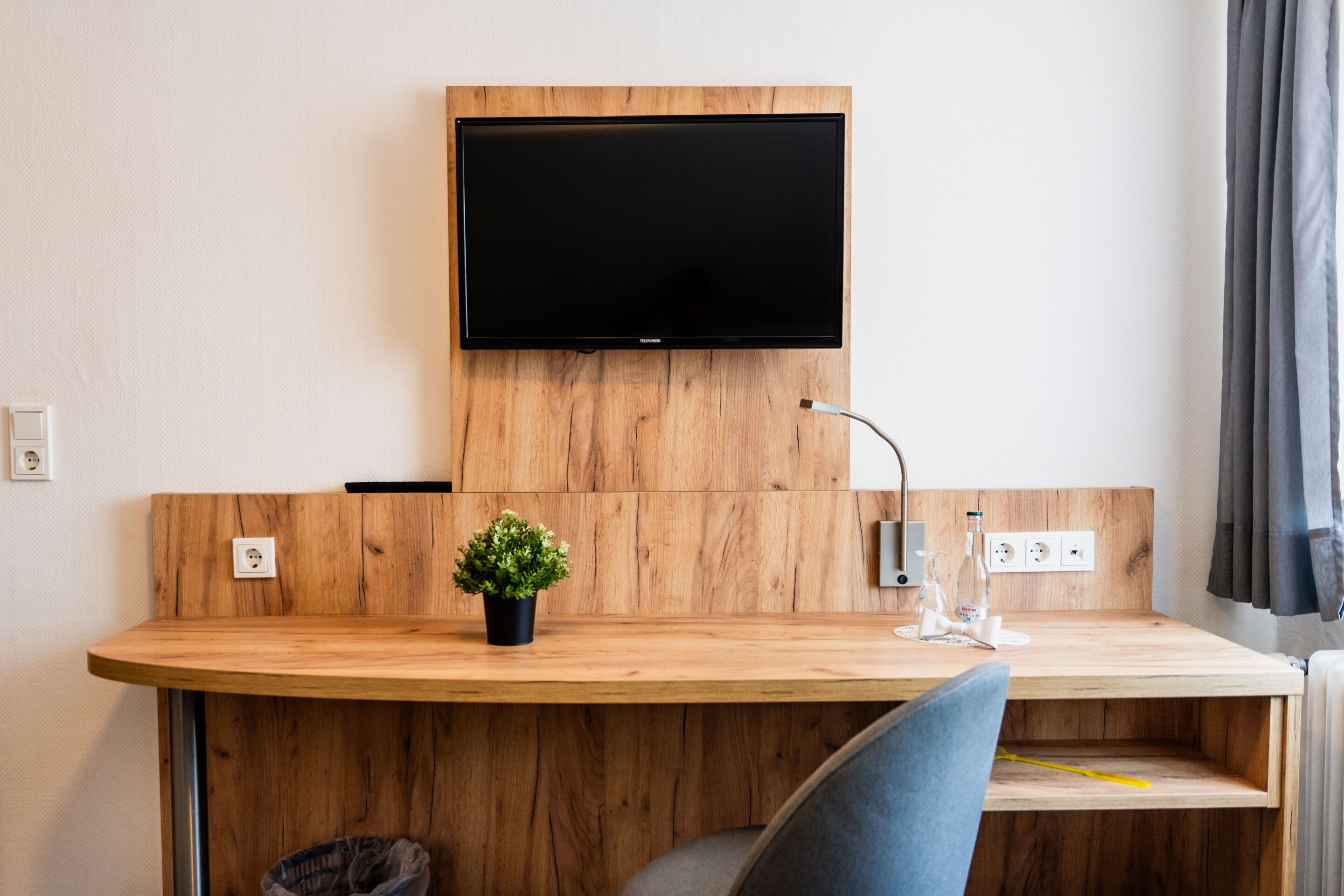 A modern room with a wooden desk, a flat-screen TV mounted above it, a small potted plant, and a bedside table with a lamp.