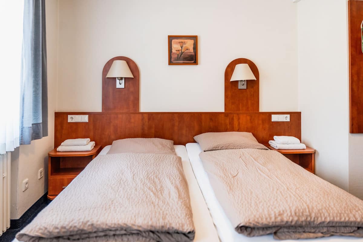 A hotel room with two single beds, wooden headboards, bedside tables with lamps, and a framed picture on the wall.