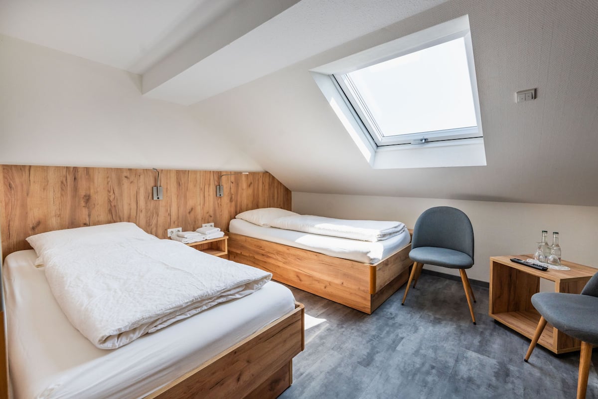 A modern bedroom with two wooden beds, a skylight, a gray chair, and a small wooden table with a remote control.