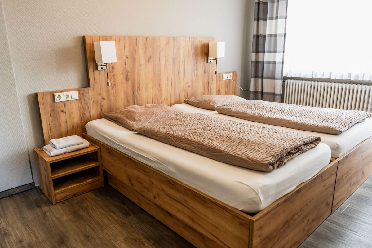 A double wooden bed with two bedside tables, each with a lamp, in a room with a radiator and a window with curtains.
