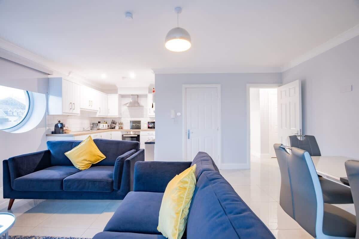 A modern living room with a blue sofa set, a dining table with chairs, and a kitchen in the background.