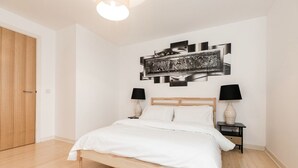 Ein Schlafzimmer mit einem hölzernen Bett, weißer Bettwäsche, zwei Nachttischen mit Lampen und einem an der Wand hängenden abstrakten Kunstwerk.