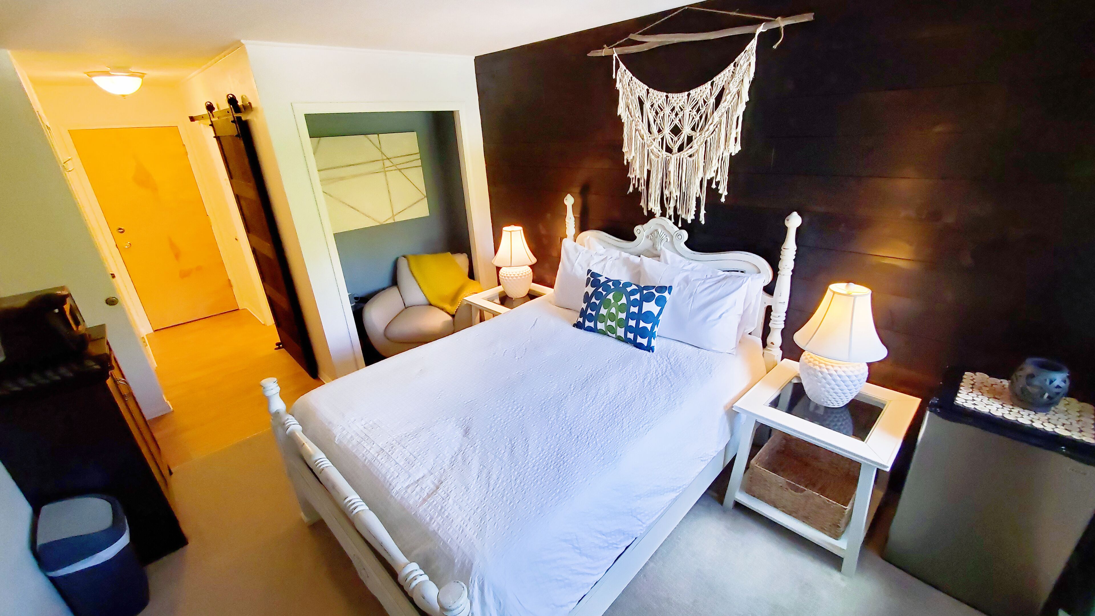 A bedroom with a bed, bedside tables, a macrame wall hanging, and a nightstand with a lamp.