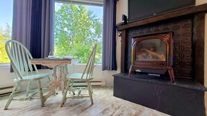 A room with a fireplace, two chairs, a table, and a window with curtains. - Open Plan Living Area, 2 Q Beds, Full Kitchen, Walk to Trails, Portable AC (Fernie)