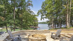 Un foyer en pierre, entouré de fauteuils Adirondack, offre une vue sur un lac avec un quai et une zone abritée en arrière-plan.