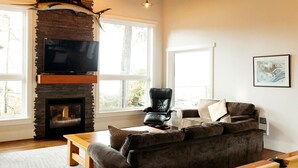 A living room with a fireplace, a leather armchair, a sofa, and a television mounted on a stone wall. - BayView House, Tofino B.C. (Tofino)
