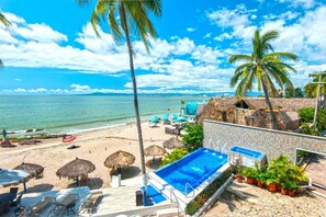 Un resort sulla spiaggia con piscina, lettini e ombrelloni di paglia.
