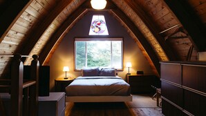 Un dormitorio acogedor en el ático con una cama, dos mesitas de noche, un aparador y un gran ventanal con vidrieras.