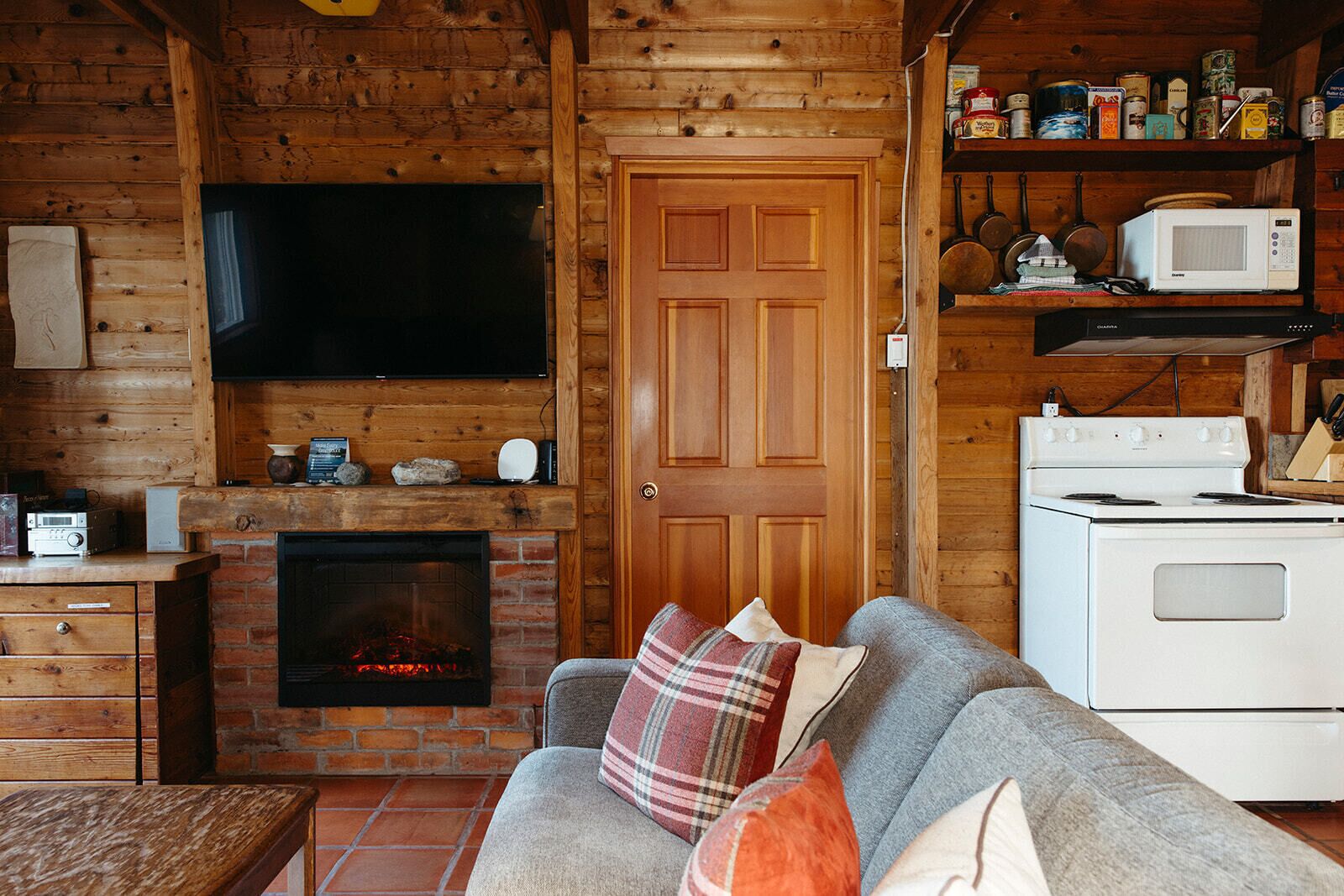 A cozy cabin interior with a fireplace, a television mounted above it, a white stove, and a sofa with plaid cushions.